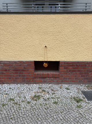 ein kinderportemonaie hängt an einem band vor einem kellerfenster. das foto wird dominiert von horizontalen linien der fassade, der gelbe putz fällt als erstes in auge, drunter ein rotes band ziegelsteine, weiter unten graues Pflaster.