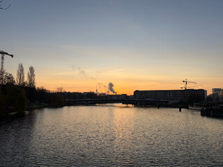 foto vom fernsehturm und einem schornstein über den nordhafen und die nordhafenbrücke hinweg. im hintergrund geht die sonne auf.