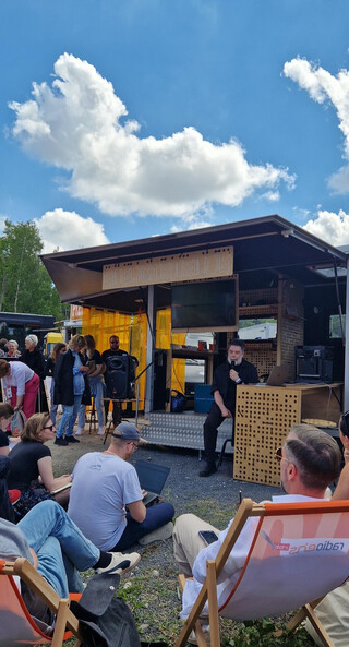 foto von der offenen bühne vom energiemobilhütte, das publikum sitzt auf gras und in orangefarbenen liegestühlen