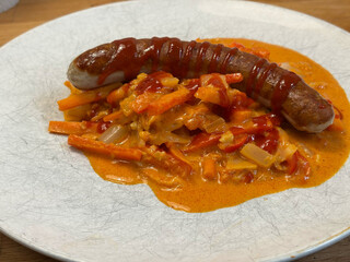 eine wurst auf rotem curry auf einem weissen teller. auf der wurst ist noch etwas siracha zu sehen