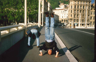 foto auf eienr brücke in lyon, peter ist von hinten zu sehen, wie er sich bückt und zwischen seinen beinen in die kamera guckt, ich mache einen kopfstand und trage hosenträger, jeans, cowboystiefel und ein jacket mit weste