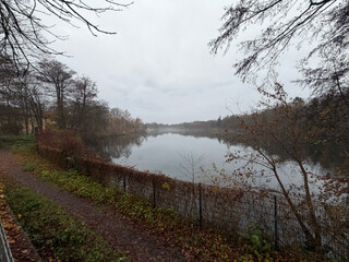 weitwinkelfoto vom plötzensee, aufgenommen am südufer. es ist diesig und grau, der see ganz ruhig, im hintergrund sieht man wasservögel.