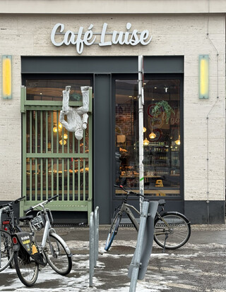 foto vom eingang des café louise. die bank vor dem eingang ist eine skulptur, eine realistische, weiss angemaclte schaumstofffigur sitzt kopfüber auf der bank in 3 meter höhe