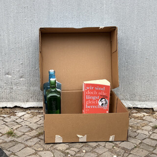 foto von einem pappkarton auf der l&uuml;tticher strasse. im karton stehen zwei leere flashcne mit kerzenst&uuml;mpfen und das buch &bdquo;Wir sind doch alle l&auml;ngst gleichberechtigt!&ldquo; von alexandra zykunov