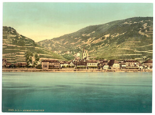 The image depicts a picturesque European village scene, likely from the late 19th to early 20th century based on its style and coloration. In the foreground is calm water that reflects the colors of the landscape above it&mdash;shades of blue and green dominate this part of the picture.