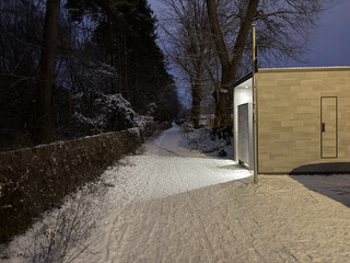 foto einer öffentlichen toilette im frühen morgengrauen. das licht aus den beiden offenen pissoir-kabinen scheint einladend auf einen schneebedeckten hohlweg