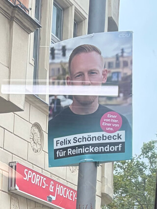 ein wahlplakat aus dem bus durchsfenstre fotografiert. auf dem plakat erkennt man, dass der kandidat seine parteizugehörigkeit (CDU) lieber nicht nennt. auf dem plakat steht nur sein name und ein slogan.