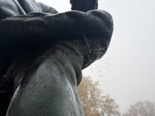 die catcherstatue an der catcherwiese in einer nahaufnahme. man sieht an der seite einer der bronzenen catcherstatuen ein spinnennetz das mit morgenreif benetzt ist.