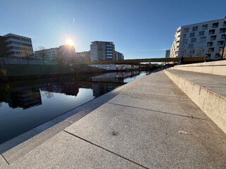 weitwinkelfoto von den stein-sitzreihen am golda-meir-steg. &uuml;ber den h&auml;usern am linken bildrand geht die sonne auf und blendet ein wenig, alles spiegelt sich im wasser des schiffahrtskanals