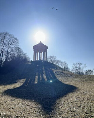 Im Gegenlicht auf einer Anhöhe ein Tempelchen, das einen riesigen Schatten auf den wintergrasigen Hang davor wirft