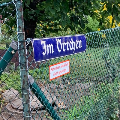 foto eines strassenschildes in einer gartensiedlung auf dem steht „im örtchen“. dadrunter ein schild mit der aufschrift „fahrräder abstellen nicht gestattet“.