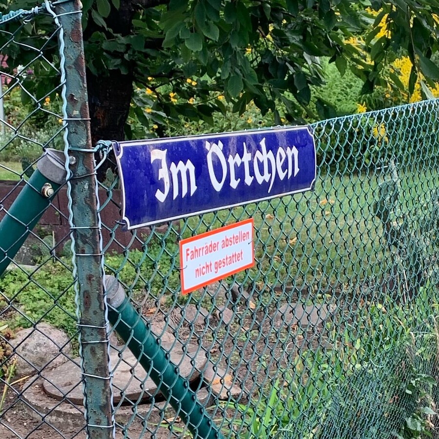 foto eines strassenschildes in einer gartensiedlung auf dem steht „im örtchen“. dadrunter ein schild mit der aufschrift „fahrräder abstellen nicht gestattet“.